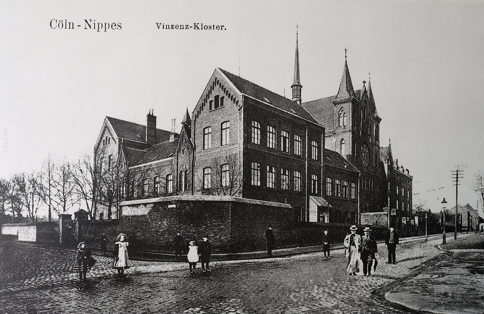 Schwarz Weiß Foto ehemaliges Vinzenz-Kloster. Heute Louise von Marillac-Schule. Alleinstehendes Backsteingebäude Neugotik Stil an der Ecke einer Kopfsteinpflaster Straße. Passanten im Kleidungsstil der 1900er-Jahre.